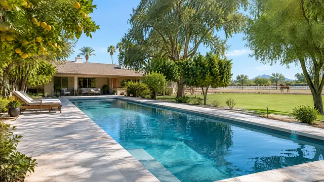 Large irrigated lot in the Lehi area of north Mesa, Arizona: a long rectangular backyard pool with a remodeled light deck, mature citrus and shade trees, a ranch-style home and an open grassy yard with a horse fence in the distance