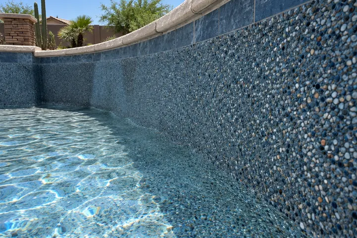 Close-up of a freshly troweled pool interior finish with exposed small pebbles in a blue-gray color, water just beginning to fill along the bottom